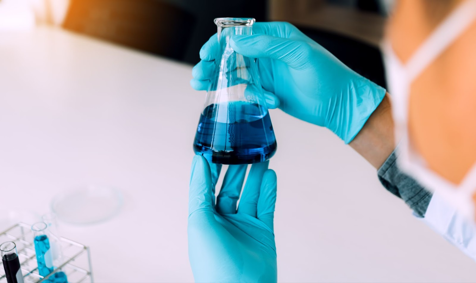 A gloved hand carefully pours blue liquid from a smaller container into a larger flask in a laboratory setting, symbolizing the precise formulation of DEF.